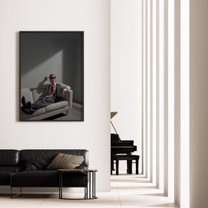 May include: A framed artwork depicts a person in a suit and sunglasses lounging on a sofa. The room features a black leather sofa, a side table, and a grand piano. The interior has a minimalist design with white walls and vertical architectural elements.
