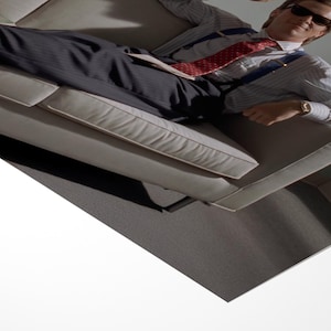 May include: A man wearing sunglasses, a striped shirt, a red patterned tie, and suspenders, is reclining on a light-colored couch. He is wearing a wristwatch. The background is a neutral gray.