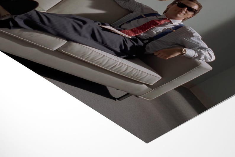 May include: A man wearing sunglasses, a striped shirt, a red patterned tie, and suspenders, is reclining on a light-colored couch. He is wearing a wristwatch. The background is a neutral gray.