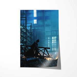May include: A poster print featuring a silhouette of Batman standing on a wooden structure, overlooking a burning scene. The background shows a large industrial building with windows, bathed in blue and white light, creating a dramatic atmosphere.