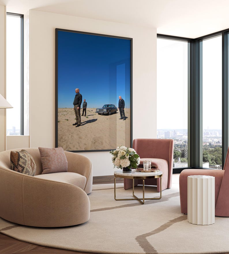 Op de afbeelding: Een moderne woonkamer met een groot ingelijst kunstwerk dat drie mannen afbeeldt die in de buurt van een klassieke auto in een woestijnlandschap staan. De kamer heeft twee gebogen, lichtbruine fauteuils, een kleine ronde tafel en een roze fauteuil.