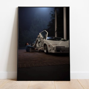 Puede incluir: Impresión enmarcada de un coche deportivo blanco con la puerta abierta. Una persona está tumbada en el suelo junto al coche. El fondo es oscuro, lo que sugiere una escena nocturna con algo de luz ambiental.