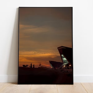 Puede incluir: Impresión artística enmarcada que representa una puesta de sol sobre un paisaje desértico con edificios modernos. El cielo transita del naranja a los tonos oscuros. Siluetas y un carro en primer plano añaden profundidad.