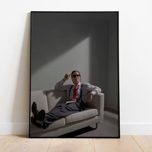 May include: Framed print featuring a man in sunglasses, seated on a gray sofa. He wears a white shirt, red tie, suspenders, and dark pants. The image is set against a gray backdrop with a beam of light.