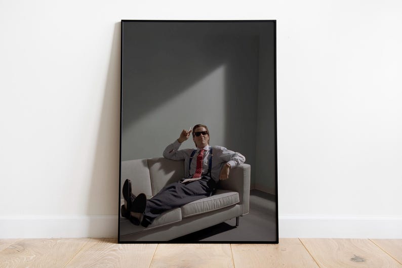 May include: Framed print featuring a man in sunglasses, seated on a gray sofa. He wears a white shirt, red tie, suspenders, and dark pants. The image is set against a gray backdrop with a beam of light.