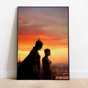 Puede incluir: P&oacute;ster enmarcado con Batman y Catwoman silueteados contra un vibrante atardecer. La obra de arte muestra a los personajes ic&oacute;nicos en una pose dram&aacute;tica, con un paisaje urbano visible en el fondo. El p&oacute;ster se exhibe sobre un suelo de madera.