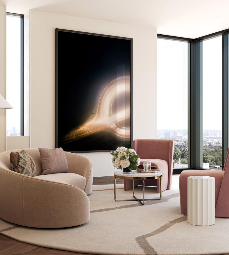 May include: A modern living room with a large framed artwork depicting a black hole. The room features two curved beige sofas with decorative pillows, two pink armchairs, a round marble coffee table, and a white side table. The room has large windows with a city view.