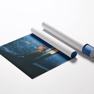 May include: A rolled-up art print with a blue and white protective tube. The print depicts a person looking out a window at night. The print is partially unrolled, revealing the image. The background is white.