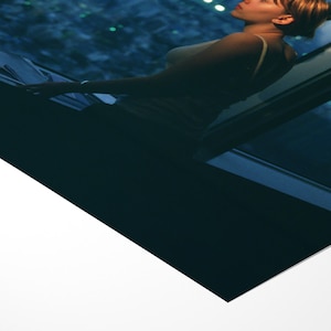 May include: A woman in a tank top stands near a window, looking up. The window frame is dark blue, and the background shows a city at night. The image has a cool color palette, with shades of blue and white.