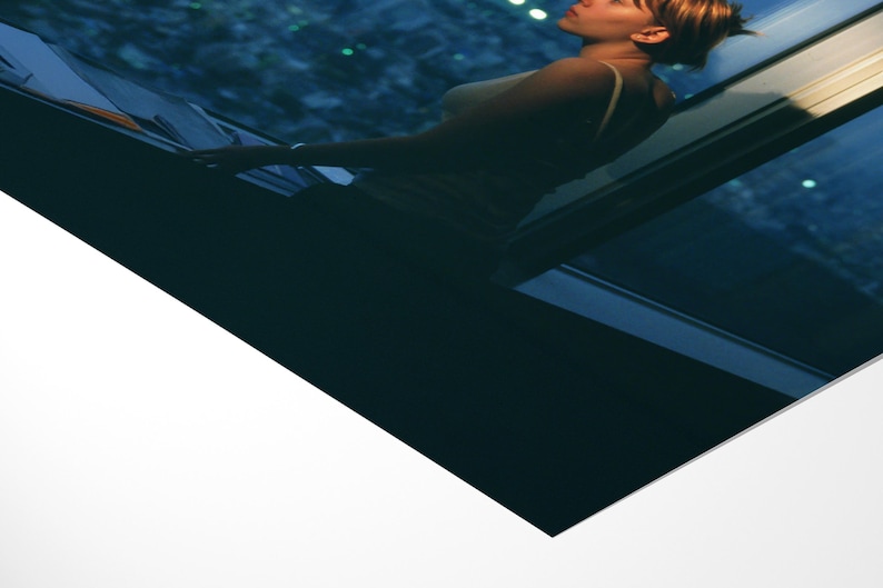 May include: A woman in a tank top stands near a window, looking up. The window frame is dark blue, and the background shows a city at night. The image has a cool color palette, with shades of blue and white.