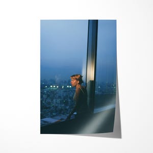 May include: A photograph of a person looking out a large window at a city skyline at dusk. The person is silhouetted against the city lights. The sky is a gradient of blues.