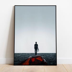 Puede incluir: Impresión artística enmarcada que muestra una persona de espaldas, silueta negra, sosteniendo un objeto rectangular. Un cielo claro y una superficie oscura se dibujan. Una forma rojo-naranja está en primer plano. La obra está sobre un suelo de madera clara.