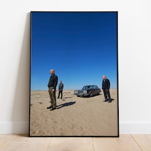 Op de afbeelding: Ingelijste kunstdruk met drie mannen in een woestijnlandschap met een klassieke auto onder een helderblauwe hemel. Het kunstwerk wordt getoond op een witte muur en een houten vloer.