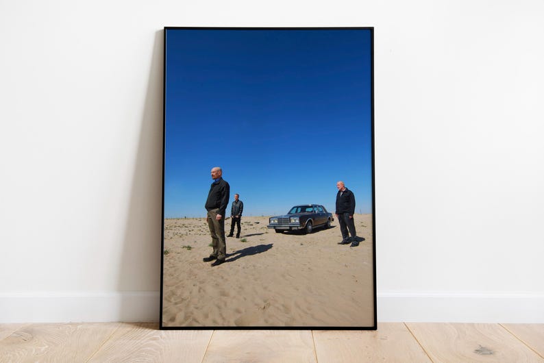 Op de afbeelding: Ingelijste kunstdruk met drie mannen in een woestijnlandschap met een klassieke auto onder een helderblauwe hemel. Het kunstwerk wordt getoond op een witte muur en een houten vloer.