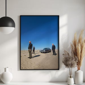 Op de afbeelding: Ingelijste kunstdruk met drie mannen in een woestijnlandschap met een klassieke auto onder een helderblauwe hemel. Het kunstwerk wordt gepresenteerd in een zwarte lijst.