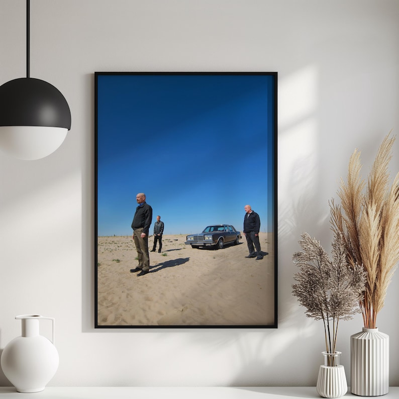Op de afbeelding: Ingelijste kunstdruk met drie mannen in een woestijnlandschap met een klassieke auto onder een helderblauwe hemel. Het kunstwerk wordt gepresenteerd in een zwarte lijst.