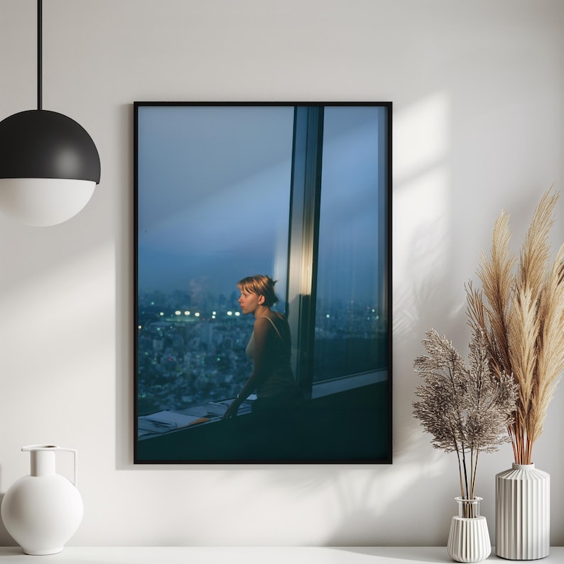 May include: A framed photograph depicts a person looking out a large window at a cityscape at dusk. The image is framed in black and is set against a white wall. Decorative vases and a hanging light are in the foreground.