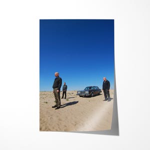 Op de afbeelding: Een posterprint met drie mannen in een woestijnlandschap, een heldere blauwe lucht en een klassieke auto. De sc&egrave;ne roept een gevoel van drama en isolement op.