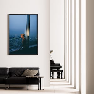 May include: A framed photograph hangs on a white wall, depicting a person looking out a window at a cityscape. A black leather sofa with throw pillows and a small side table are in the foreground. A black piano is visible in the background.