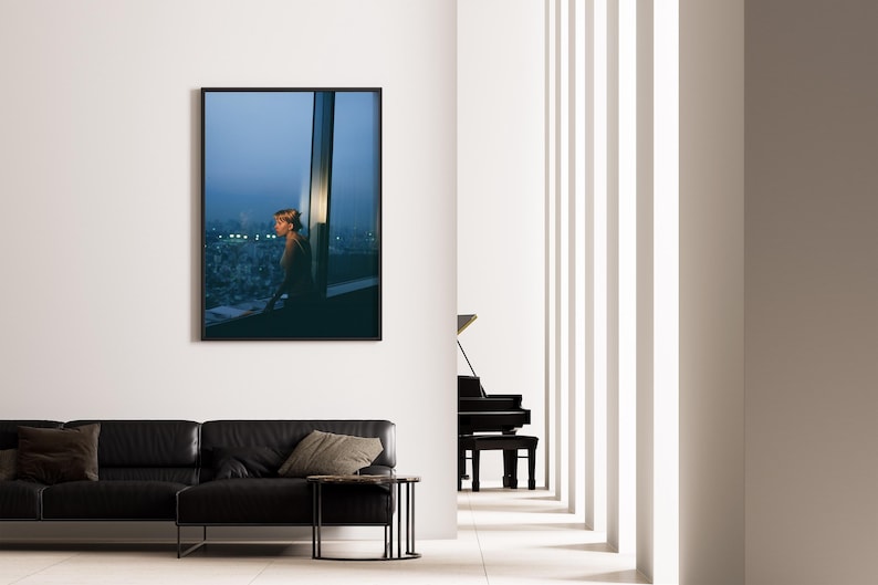 May include: A framed photograph hangs on a white wall, depicting a person looking out a window at a cityscape. A black leather sofa with throw pillows and a small side table are in the foreground. A black piano is visible in the background.