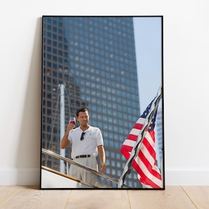 Puede incluir: Impresión enmarcada de un hombre sosteniendo una copa de vino tinto, con una bandera estadounidense de fondo. El hombre lleva un polo blanco y pantalones color caqui. Se ve un edificio alto.