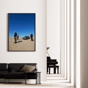 Op de afbeelding: Een ingelijste foto toont drie personen bij een klassieke auto in een woestijnlandschap onder een helderblauwe hemel. De foto hangt aan een witte muur boven een zwarte leren bank.