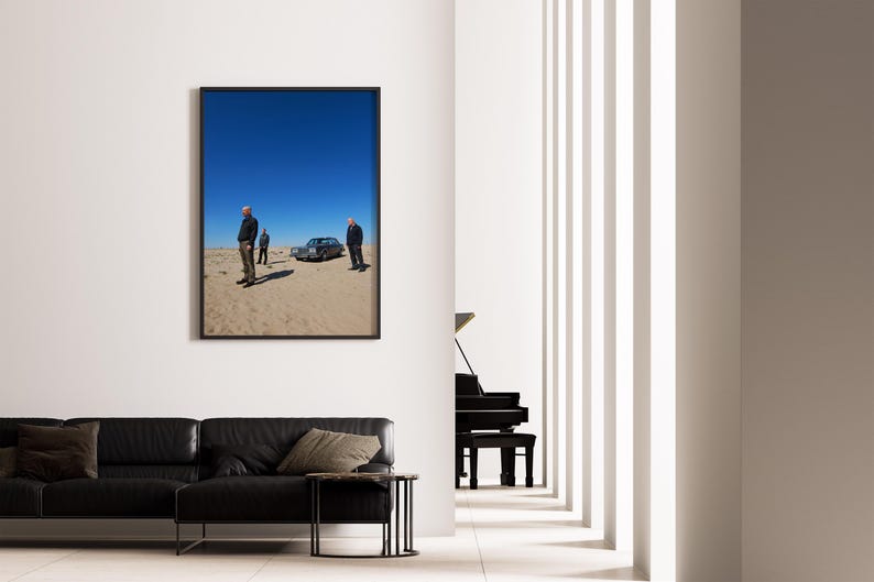 Op de afbeelding: Een ingelijste foto toont drie personen bij een klassieke auto in een woestijnlandschap onder een helderblauwe hemel. De foto hangt aan een witte muur boven een zwarte leren bank.