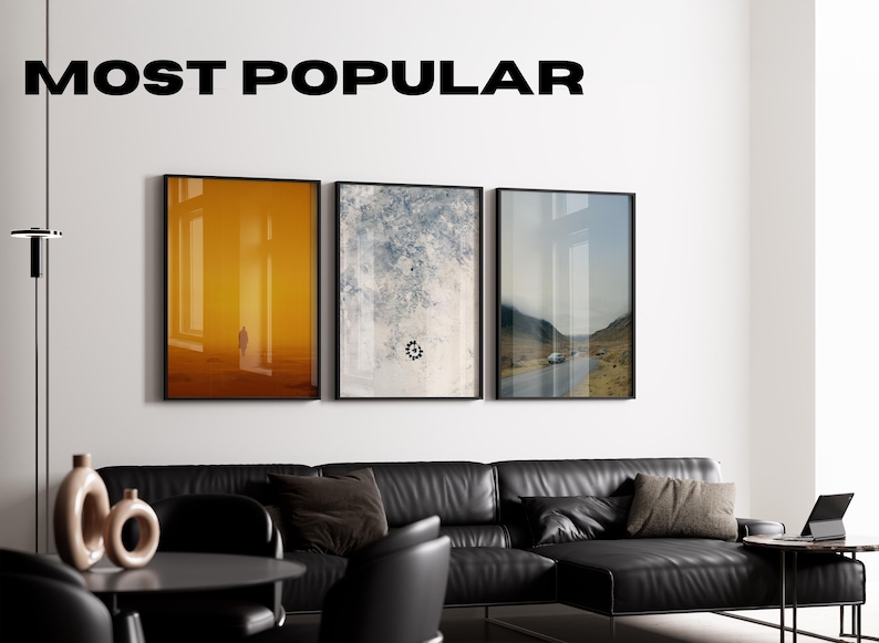 May include: Three framed art prints hang above a black leather sectional sofa. The prints feature abstract and landscape scenes. The text "MOST POPULAR" is displayed above the artwork. A black table, chairs, and a floor lamp complete the modern living room setting.