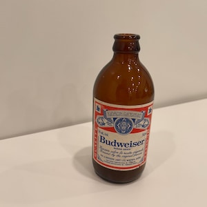 Puede incluir: Una botella de cerveza Budweiser de vidrio marrón con una etiqueta roja, blanca y azul. La etiqueta incluye el logotipo y el texto de Budweiser. La botella está vacía y parece ser un artículo vintage.