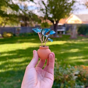 Könnte beinhalten: Ein kleiner Terrakotta-Topf enthält eine Miniaturpflanze mit blauen Glasblättern und Kupferdrahtstielen. Die Pflanze wird vor einem verschwommenen grünen Hintergrund gehalten, der eine Umgebung im Freien suggeriert. Der Topf hat einen Durchmesser von etwa 4 cm.