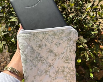 Op de afbeelding: Een zwarte e-reader met het Amazon-logo wordt vastgehouden in een hoesje met bloemenprint. De hoes is wit met een groen bloemenpatroon en een witte rits. De e-reader is gedeeltelijk zichtbaar, wat suggereert dat deze erin wordt opgeborgen.