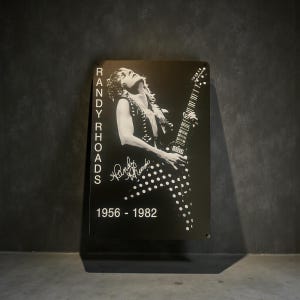 May include: Black and white metal sign featuring a portrait of Randy Rhoads playing a guitar. The sign includes the text "RANDY RHOADS" vertically on the left side, his signature, and the years "1956 - 1982".
