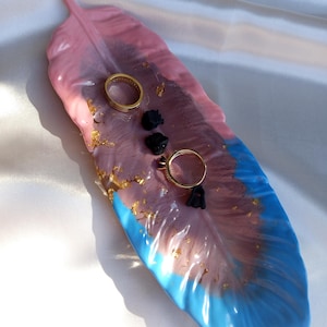 May include: A feather-shaped resin tray in pink, purple, and blue, with gold leaf accents. Two gold rings and three small black decorative objects rest on the tray. The tray is designed for jewelry or small items.