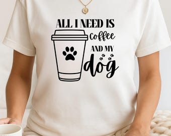 May include: Cream-colored t-shirt with the text "ALL I NEED IS coffee AND MY dog" in black. A black graphic of a coffee cup with a paw print and paw prints is also on the shirt. A gold necklace is visible.