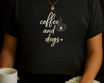 May include: Black t-shirt with the words "coffee and dogs" in a cursive font. A coffee cup with a paw print is next to the text. The person is wearing a gold necklace and holding a white mug.