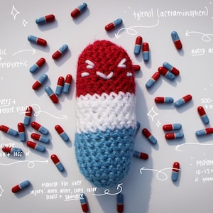 May include: A crocheted pill in red, white, and blue with a smiling face, surrounded by red and blue capsules. Text includes "analgesic", "antipyretic", and "tylenol". The image is a whimsical representation of medication.