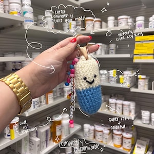 May include: A handmade, crocheted pill-shaped keychain in cream and blue, with a smiling face. The keychain has a beaded strand with the text "TAKE UR MEDS". The background shows pharmacy shelves with various medications.