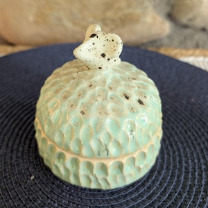 Peut inclure: Un petit pot en céramique vert clair avec un couvercle, présentant une surface texturée et alvéolée avec des taches brun foncé. Le couvercle est surmonté d'une poignée sculptée en forme de souris tachetée, ajoutant un détail décoratif.