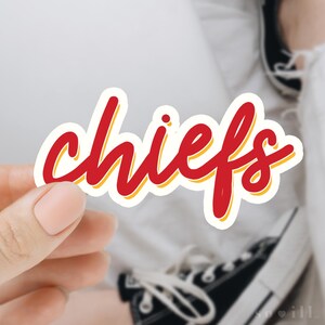 Puede incluir: Una pegatina "chiefs" roja y amarilla con un contorno blanco. La pegatina est&aacute; siendo sostenida por una mano que lleva un anillo.