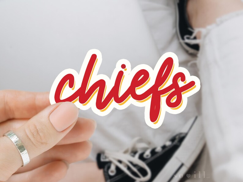 Puede incluir: Una pegatina "chiefs" roja y amarilla con un contorno blanco. La pegatina est&aacute; siendo sostenida por una mano que lleva un anillo.
