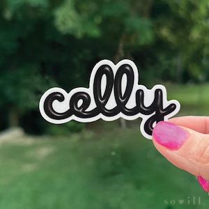 May include: A white sticker with the word "celly" in black, cursive lettering. The sticker is held up against a blurred green background. The word "so ill" is printed in the bottom right corner.