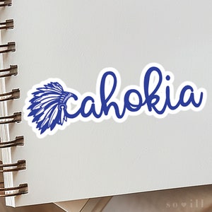 May include: Blue and white sticker with the word "Cahokia" written in a cursive font. A blue headdress with feathers is to the left of the word.