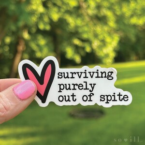 May include: A white sticker with the text "surviving purely out of spite" in black font. A pink heart with a black outline is on the left. The sticker is held up by a hand with pink nail polish.