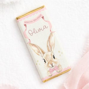 May include: A chocolate bar with gold foil wrapping. The front features a watercolour illustration of a bunny wearing a pink bow tie. The text "Special Delivery" and the name "Olivia" are displayed in pink script on a banner.