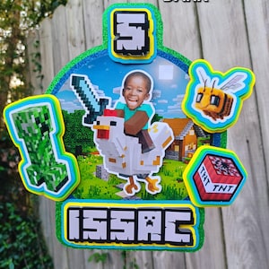 May include: A colorful Minecraft-themed cake topper featuring a young boy riding a pixelated chicken, holding a sword. The topper includes the name "Isaac" and the number "5", along with Minecraft characters like a Creeper, bee, and TNT block. The text "SKIN COMPLEXION: DARK" is also visible.