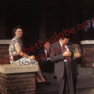 May include: A group of people standing on a porch, one person is holding a raccoon. The woman on the left is wearing a floral dress and the man on the right is wearing a white shirt and a dark suit.