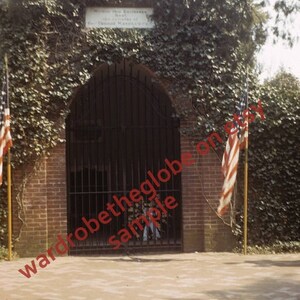 May include: A brick building with a black metal gate and a sign that reads "Within this Enclosure Rest the Remains of GEORGE WASHINGTON PRESIDENT OF THE UNITED STATES". Two American flags are on poles in front of the building.