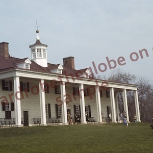 May include: Exterior view of a large, white colonial-style building with a dark brown roof and a cupola. The building has a long porch supported by white columns. A person in a coat and hat stands in the foreground.