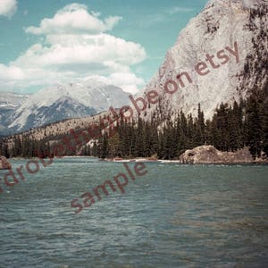 May include: A scenic view of a river flowing through a mountain valley. The river is calm and the water is a deep blue-green color. The mountains are covered in trees and the sky is a bright blue with white clouds.