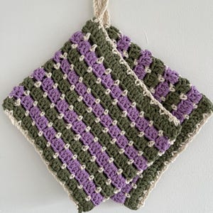 May include: Handmade crochet potholder in a square shape. The potholder features a checkerboard pattern of olive green and lavender purple squares, with off-white accents and edging. A loop is attached for hanging.